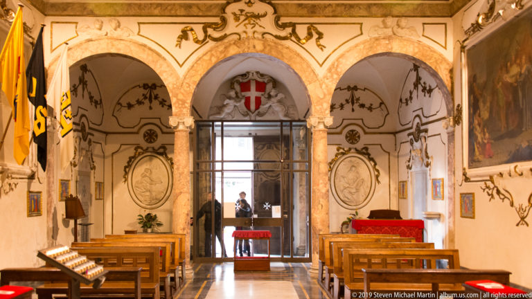Small decorated chapel with marble columns, wooden benches, flags and a red shield bearing a white eight-pointed cross.
