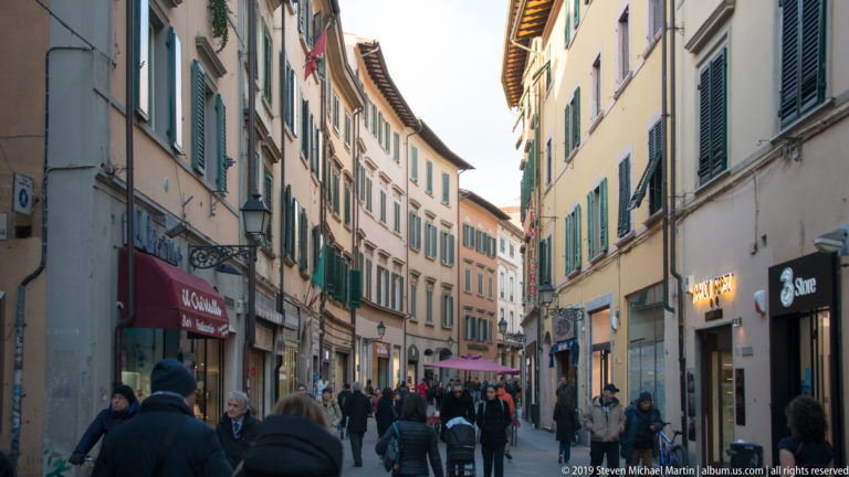 Crowded narrow shopping street lined with pastel buildings, shutters, storefronts and pedestrians in central Pisa.