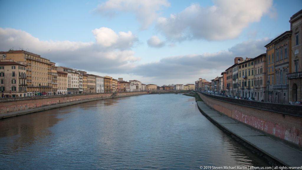 Arno River flowing between embankment walls and rows of historic Pisan buildings under a partly cloudy sky.