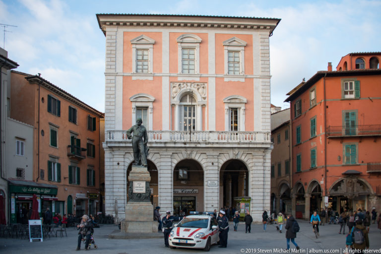 Statue of Giuseppe Garibaldi before a pink-and-white palazzo with arcades, pedestrians and street activity in Piazza Garibaldi.