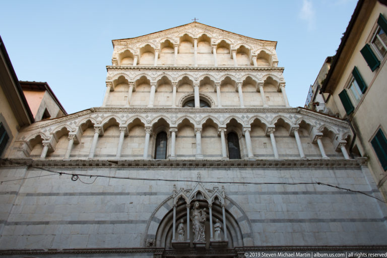 Upward view of San Michele in Borgo with tiered arcades, slender columns and a pointed portal between neighboring buildings.