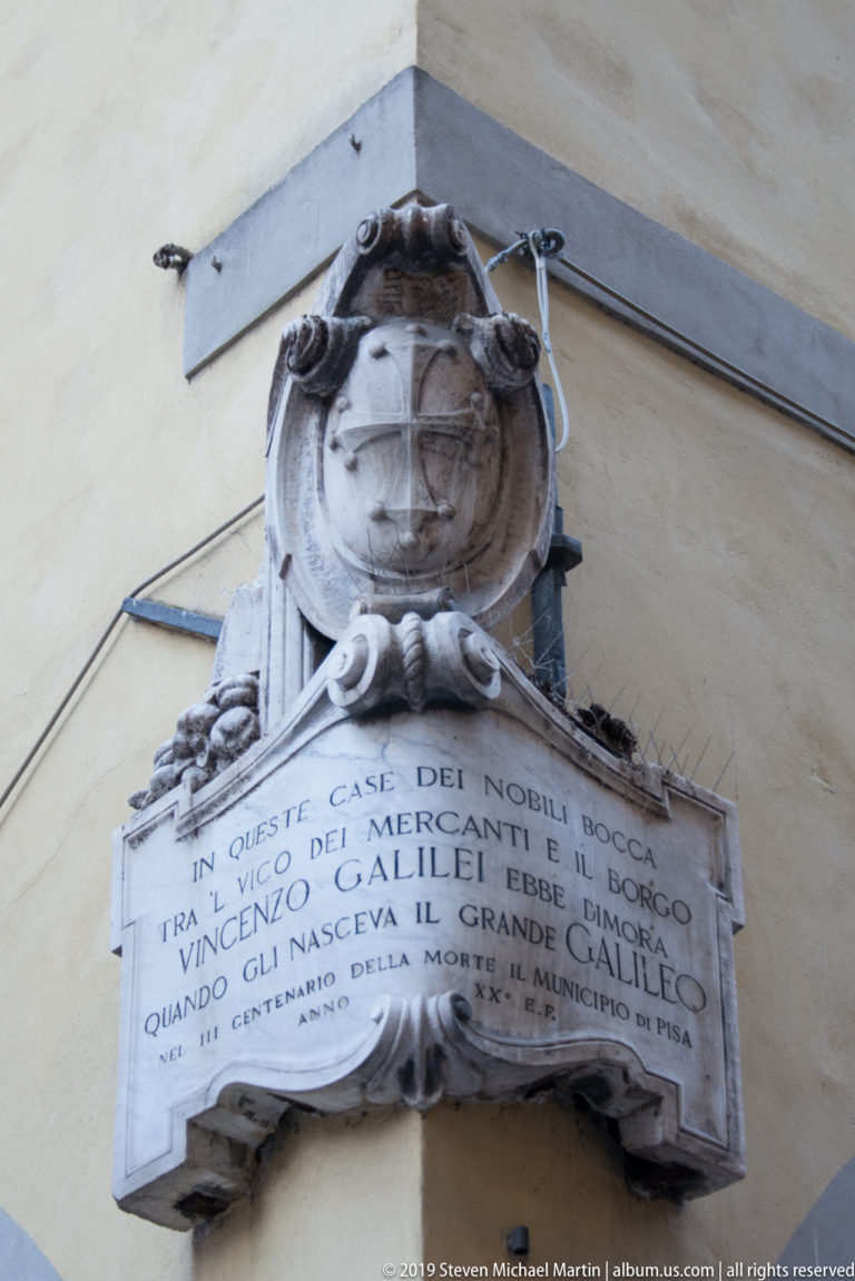 Marble Italian inscription about Galileo Galilei beneath a weathered sculpted crest on a stucco wall.