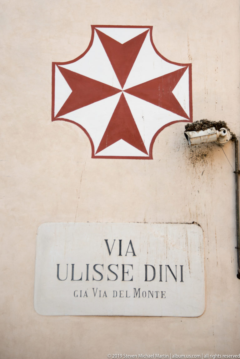 Red eight-pointed cross painted above a marble street sign reading Via Ulisse Dini, già Via del Monte.