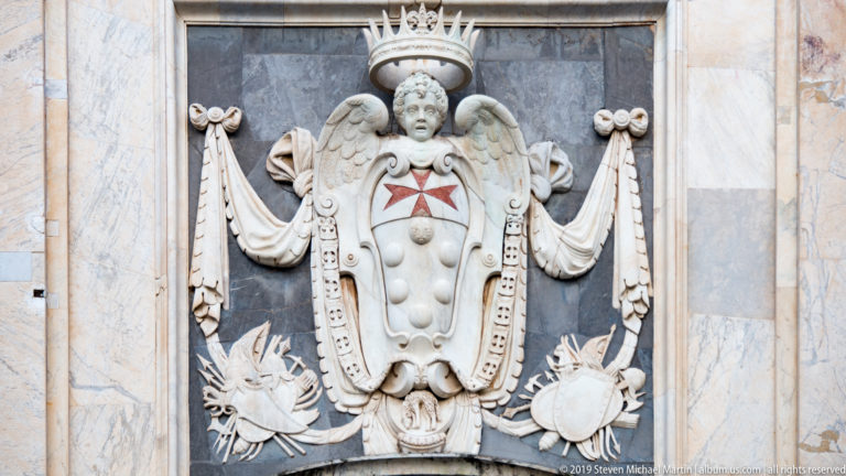 Crowned winged figure above a white marble shield with red cross, drapery and radiating ornaments on a dark panel.