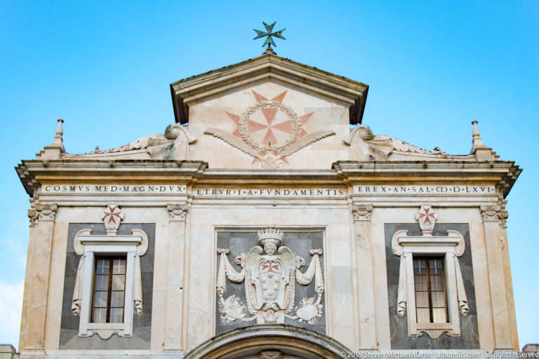 Upper church façade with Latin inscription, paired windows, carved shield and multiple red cross emblems beneath the pediment.