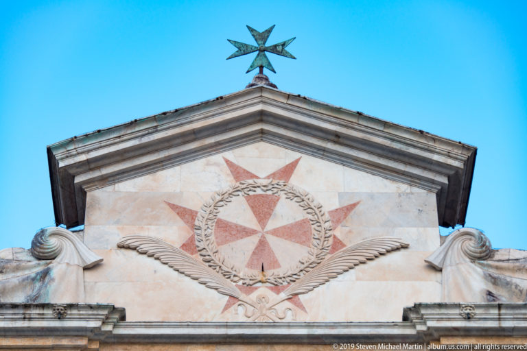 Triangular church pediment bearing an eight-pointed red cross in a wreath, topped by a metal finial.