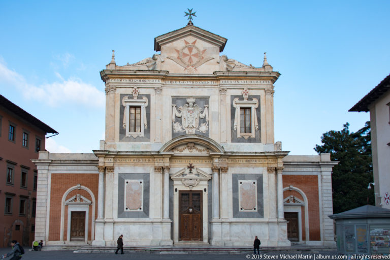 Front façade of Santo Stefano dei Cavalieri with marble center, brick side bays and cross emblems beneath a blue sky.