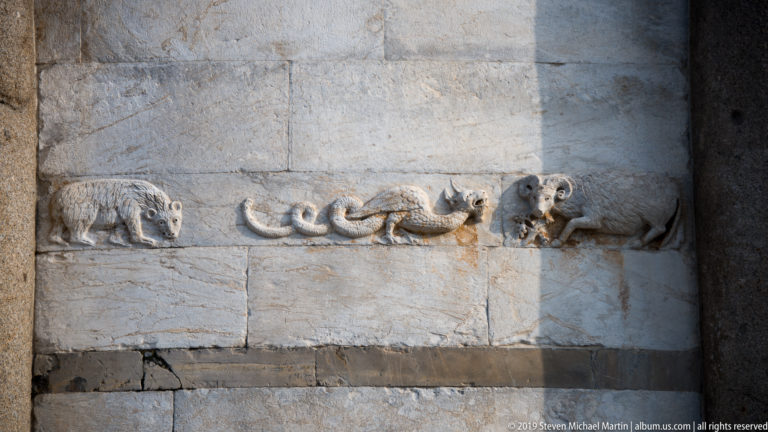 Weathered animal reliefs, including quadrupeds and a long serpent-like creature, carved across pale marble blocks.