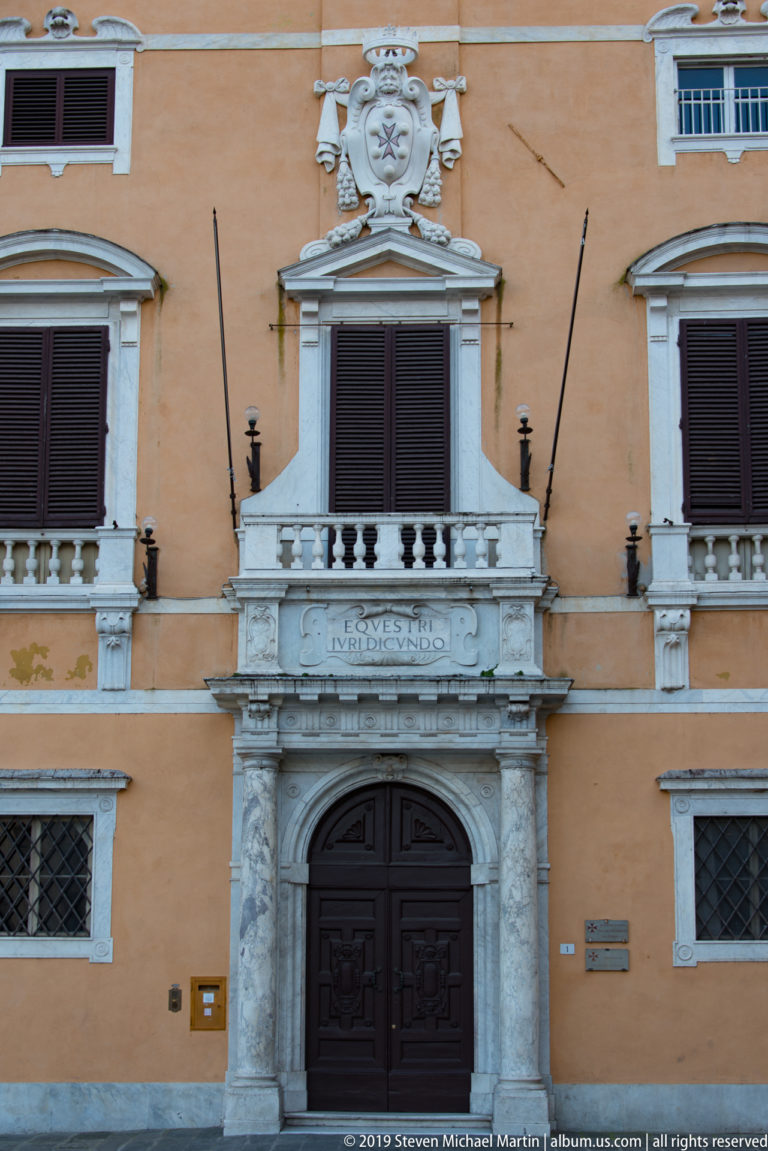 Marble-framed doorway with dark wooden doors, inscribed balcony and heraldic ornament on an ochre palazzo façade.