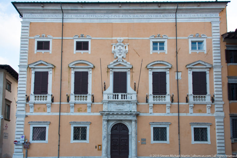 Symmetrical ochre palazzo with dark shutters, pale stone trim, central marble doorway and heraldic relief above.