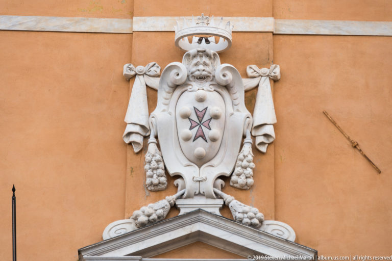 White stone crowned coat of arms with a cross, scrolls and drapery mounted on an ochre wall above a pediment.