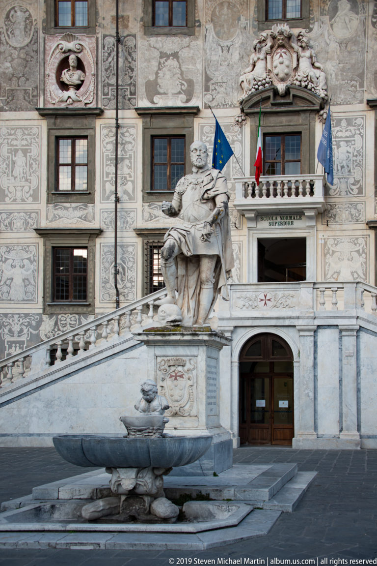 Stone fountain and seated monument before the staircase, flags and decorated façade of Palazzo della Carovana.