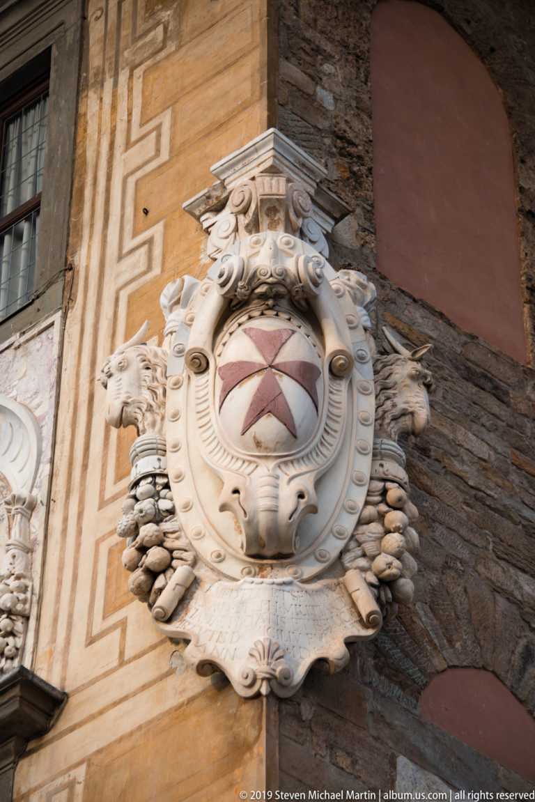 Ornate stone cartouche with an eight-pointed Maltese cross carved in relief on a historic wall in Pisa.