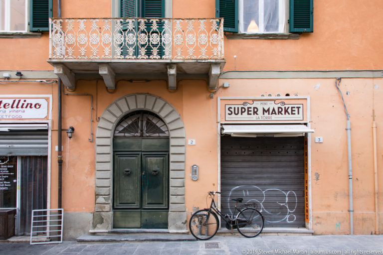 Closed Super Market storefront, bicycle, arched doorway, balcony, and green shutters on a peach building in Pisa.