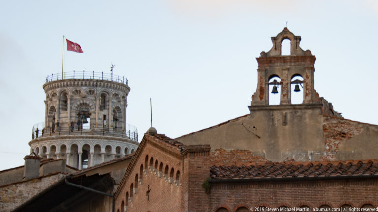 Upper Leaning Tower rises behind terracotta roofs and brick walls beside a small two-bell gable.