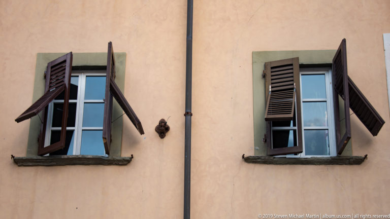 Two shuttered windows with stone surrounds on a peach stucco wall, divided by a dark vertical drainpipe.