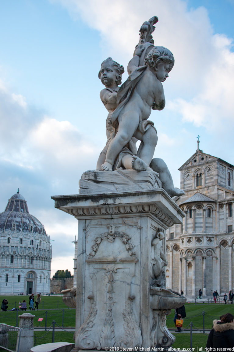 Side view of the Fontana dei Putti, with twisting marble figures and Pisa’s Baptistery and Cathedral behind.