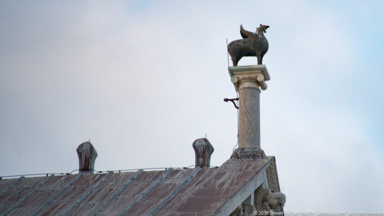 Small dark animal sculpture mounted on a pale stone column above a weathered tiled roof in Pisa.