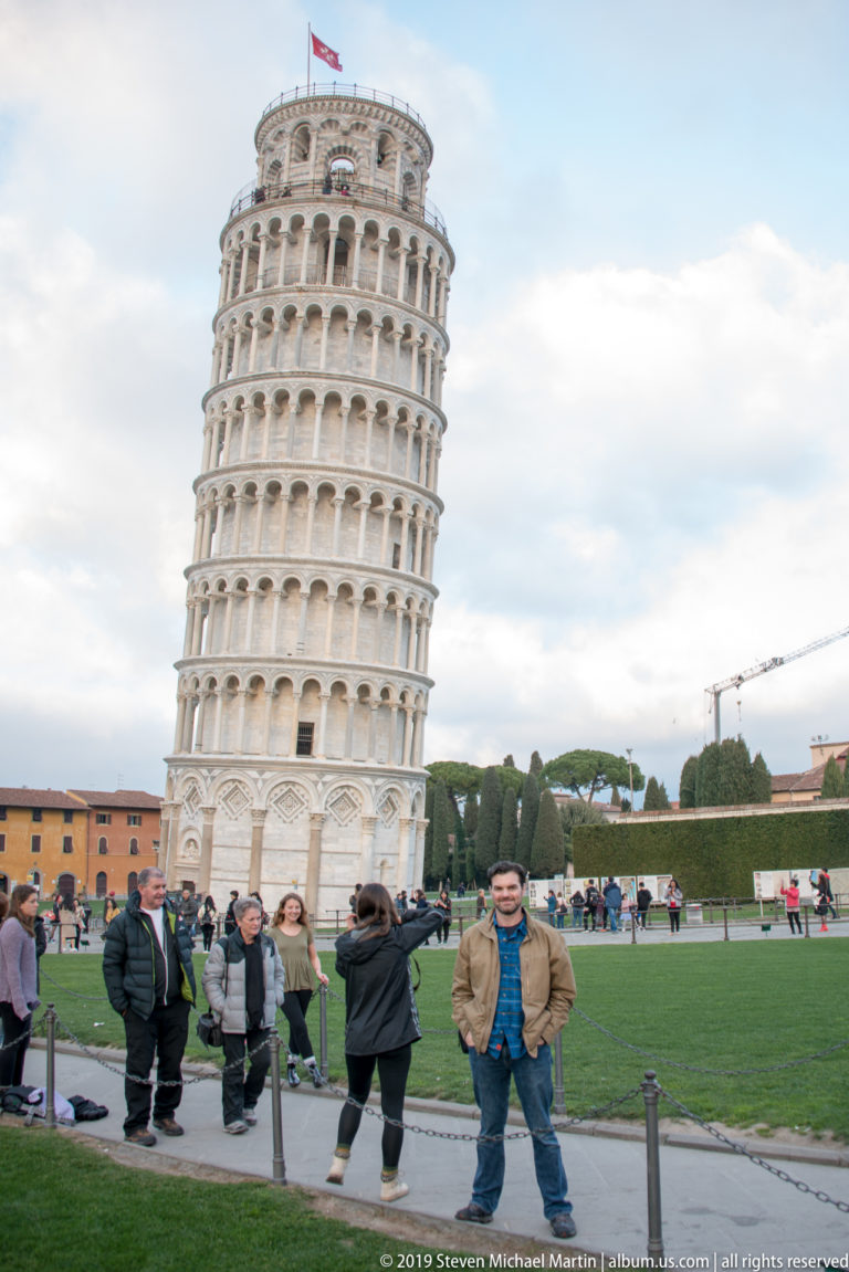Leaning Tower behind a busy group of visitors, with a man posed near the edge of the Piazza dei Miracoli lawn.