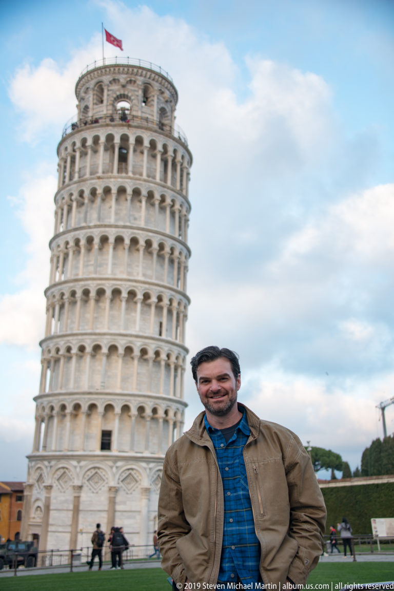 Man in brown jacket posed before the Leaning Tower, with lawn and visitors visible around its base.