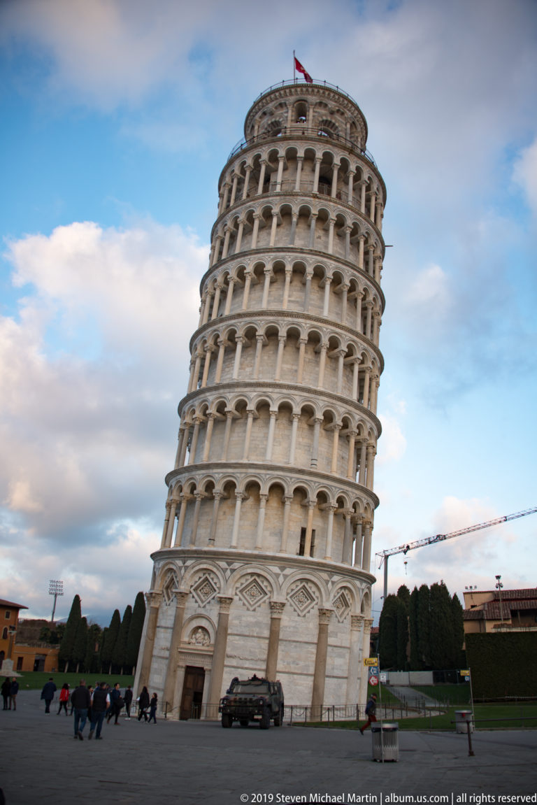 Leaning Tower of Pisa at evening, with visitors at its base, clipped hedges, and a crane in the distance.