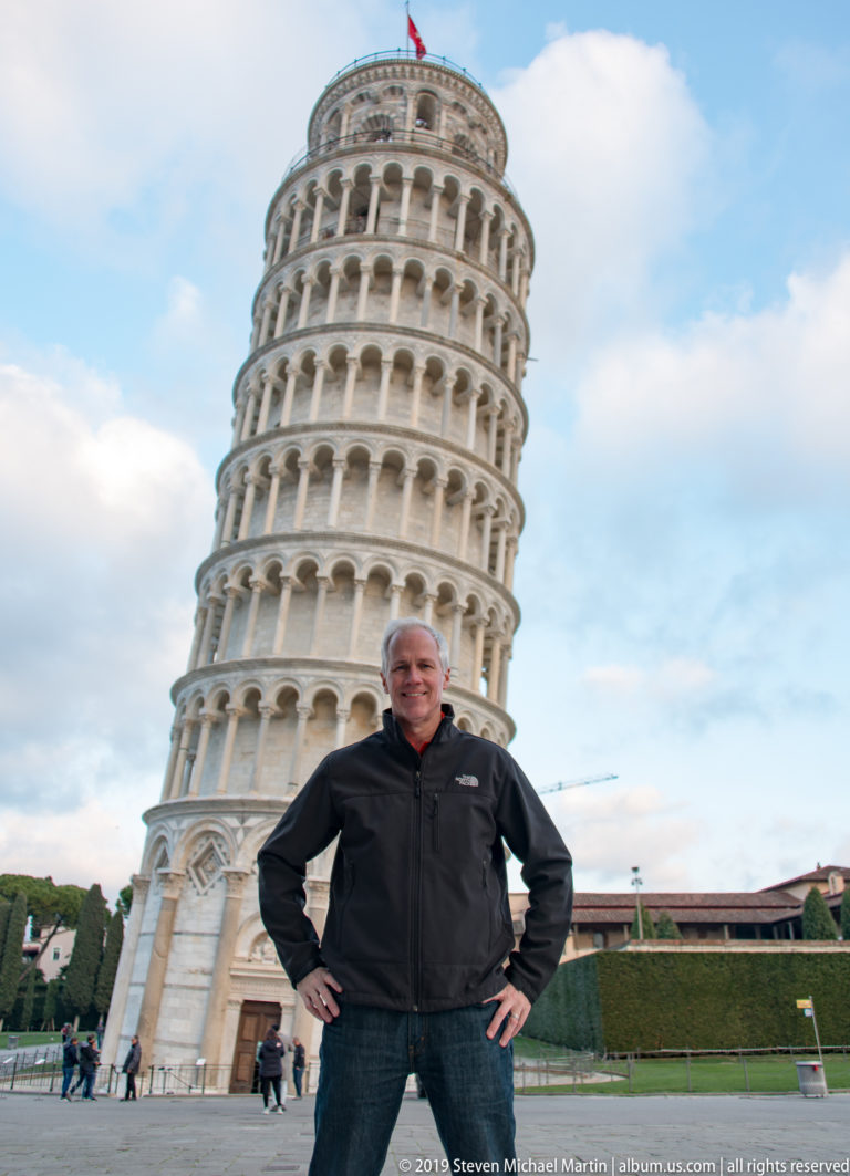 Man stands hands-on-hips on the Piazza dei Miracoli with the full Leaning Tower directly behind him.