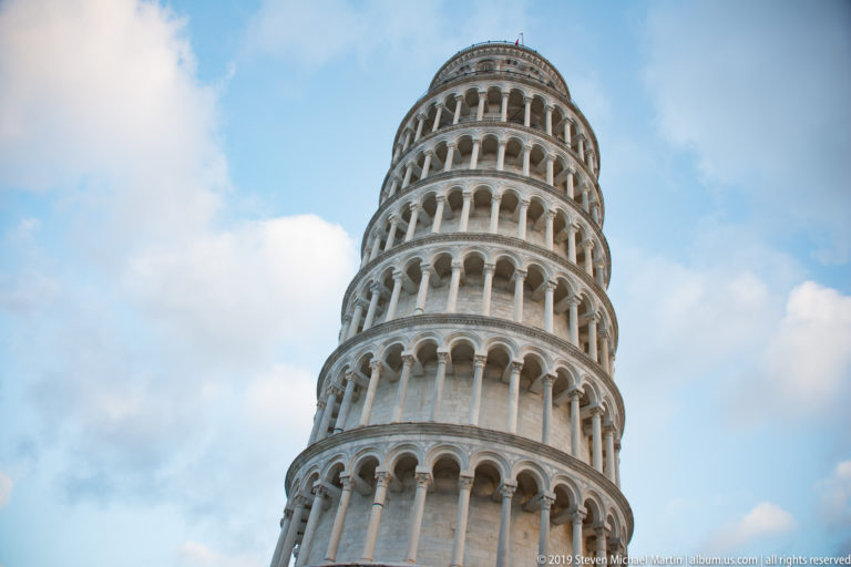 Upper arcaded stories of the Leaning Tower of Pisa rise against pale blue sky and clouds.