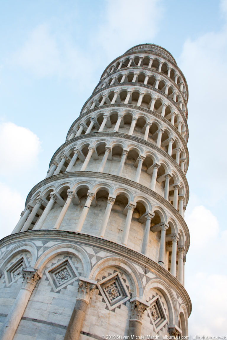 Steep upward view from the base of the Leaning Tower, with marble arcades curving diagonally toward the sky.