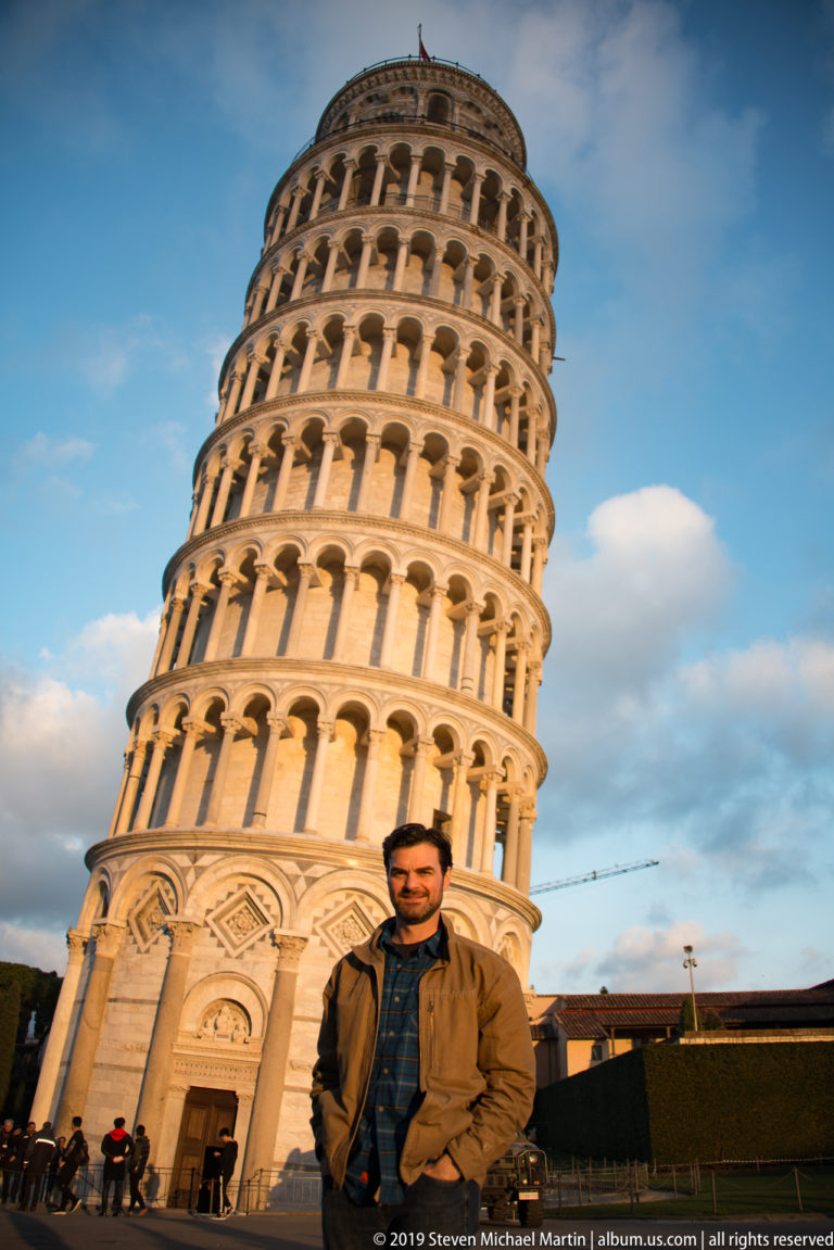 Man posed at the base of the Leaning Tower, whose tilted arcades fill most of the background.