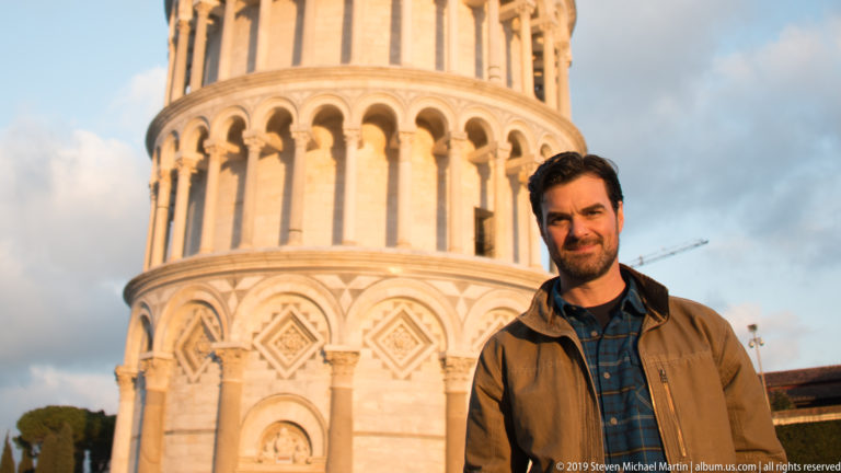 Man in brown jacket stands before the sunlit lower marble arcades of the Leaning Tower of Pisa.