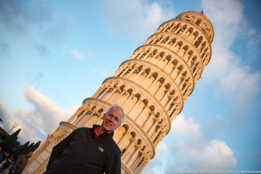 Man in dark jacket posed close to camera with the Leaning Tower of Pisa rising diagonally behind him.