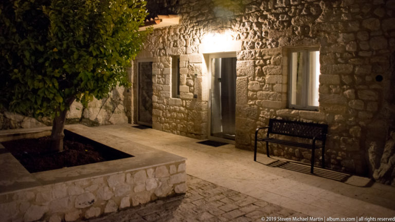 Rustic stone façade with doorway, shuttered window, metal bench, and leafy tree growing from a square courtyard planter.