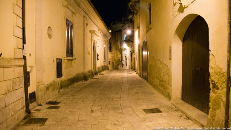 Narrow stone-paved lane between historic buildings, with a tall arched wooden doorway and wall lamps receding into the distance.