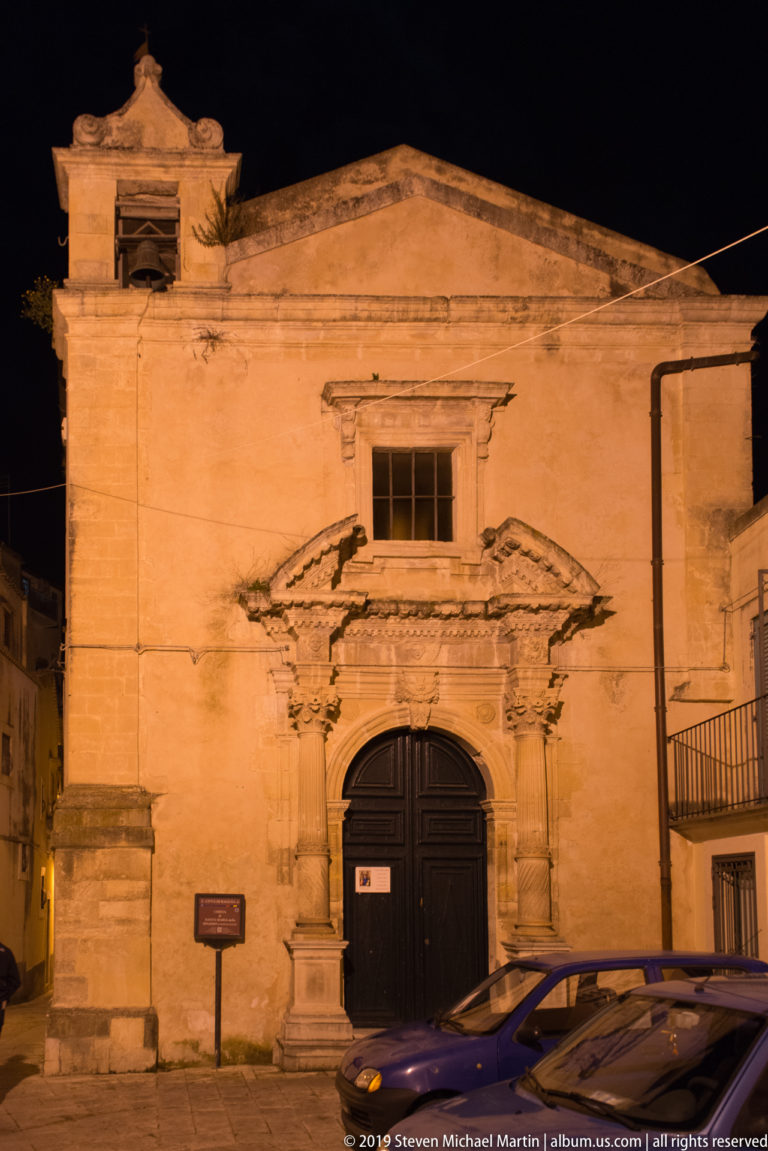 Warmly lit gabled church façade with dark arched door, elaborate stone entrance surround, upper window, and parked cars.