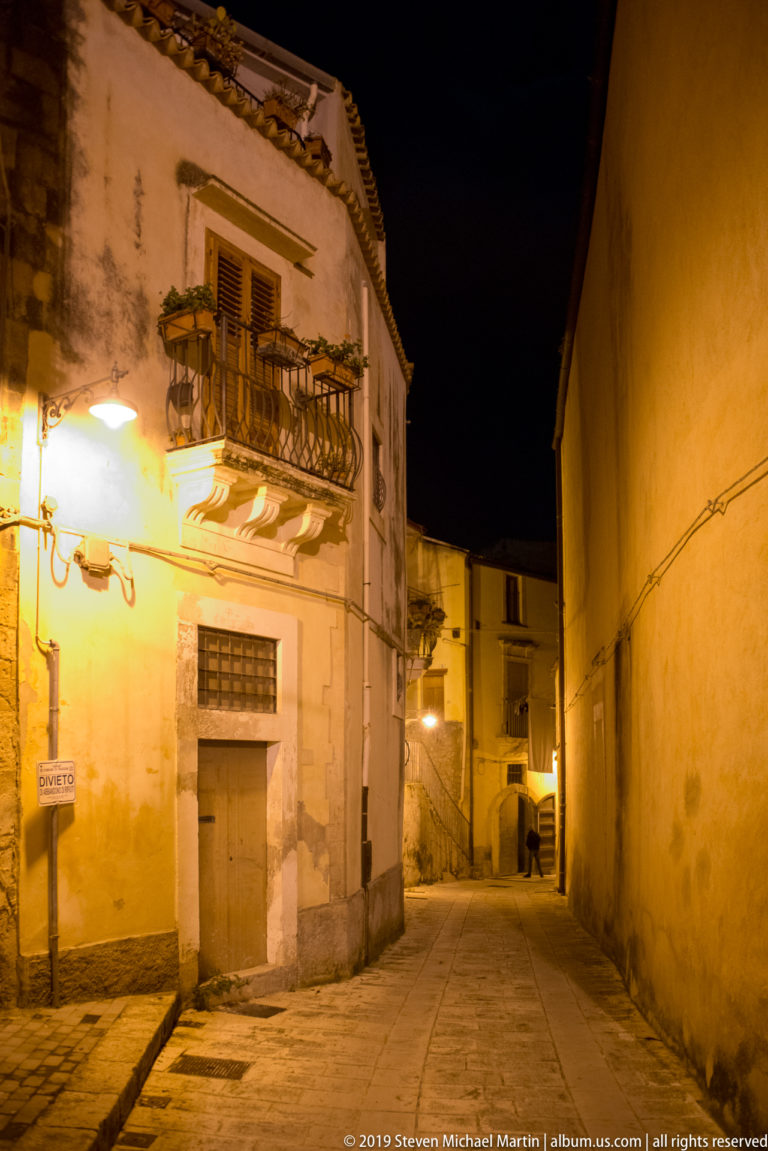 Night street corner with weathered house, projecting iron balcony on stone brackets, shuttered openings, and a narrow lane beyond.
