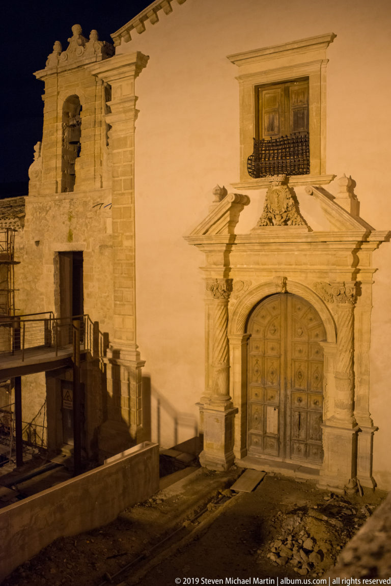 Small illuminated church façade with carved stone portal, paneled wooden doors, curving pediment, upper window, and iron balcony.