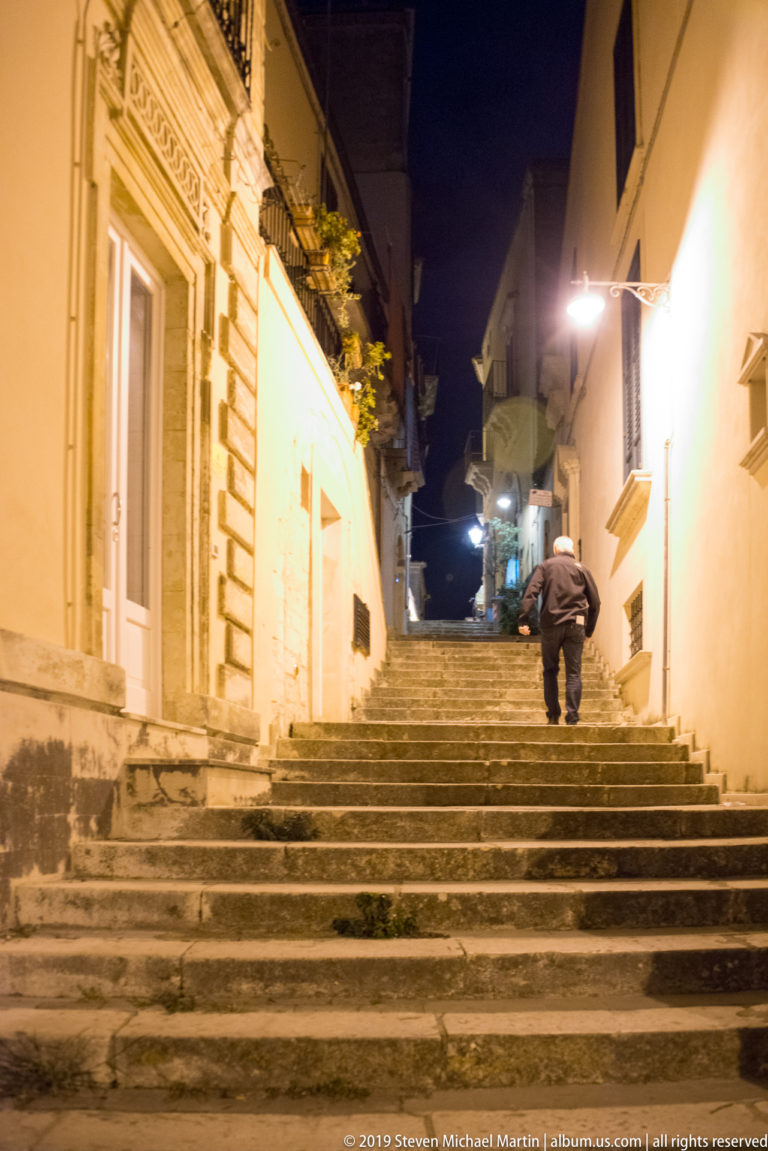 Pedestrian climbing broad stone steps between closely spaced buildings under bright streetlights at night.