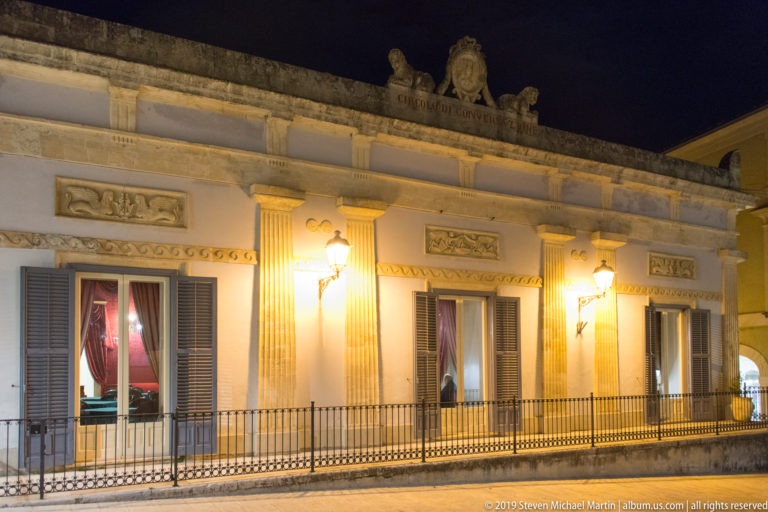 Illuminated pale residential façade with iron balcony, shuttered French doors, fluted pilasters, relief panels, and red curtains.