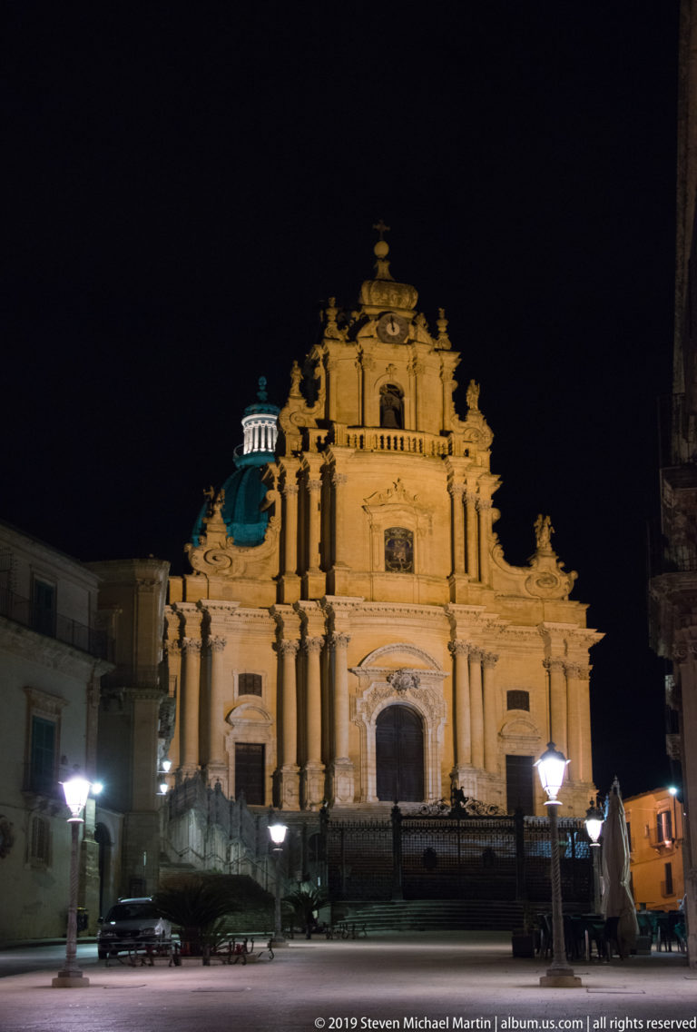Frontal night view of the illuminated Duomo di San Giorgio across Piazza Duomo, with streetlamps and parked vehicles in front.
