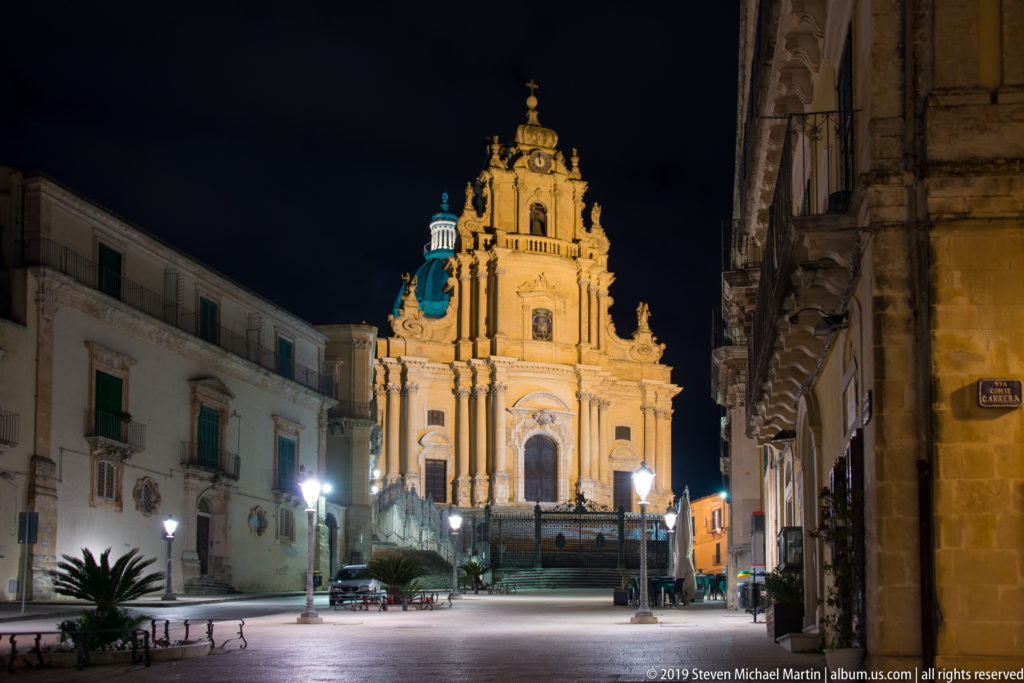 Duomo di San Giorgio illuminated at the end of an open square, framed by historic buildings, streetlamps, cars, and a small palm.