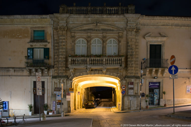 Historic stone building spanning a brightly lit street arch, with ornate upper windows, balcony, surrounding façades, and a car beneath.