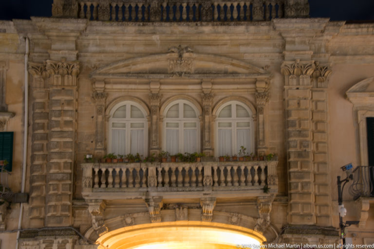 Ornate stone façade with three arched windows, balustraded balcony, rusticated pilasters, and a brightly lit arch below.