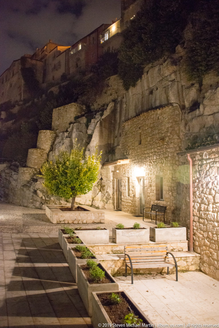 Night courtyard with pale stone terraces, benches, planting beds, masonry buildings, and a rugged rock face rising behind.