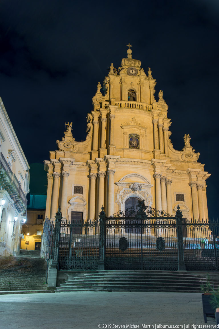 Illuminated Baroque façade of the Duomo di San Giorgio above broad steps and an iron fence in Ragusa Ibla at night.