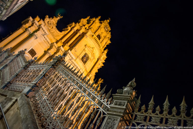 Low-angle view of a floodlit Baroque church façade rising behind decorative iron fencing and pointed finials at night.