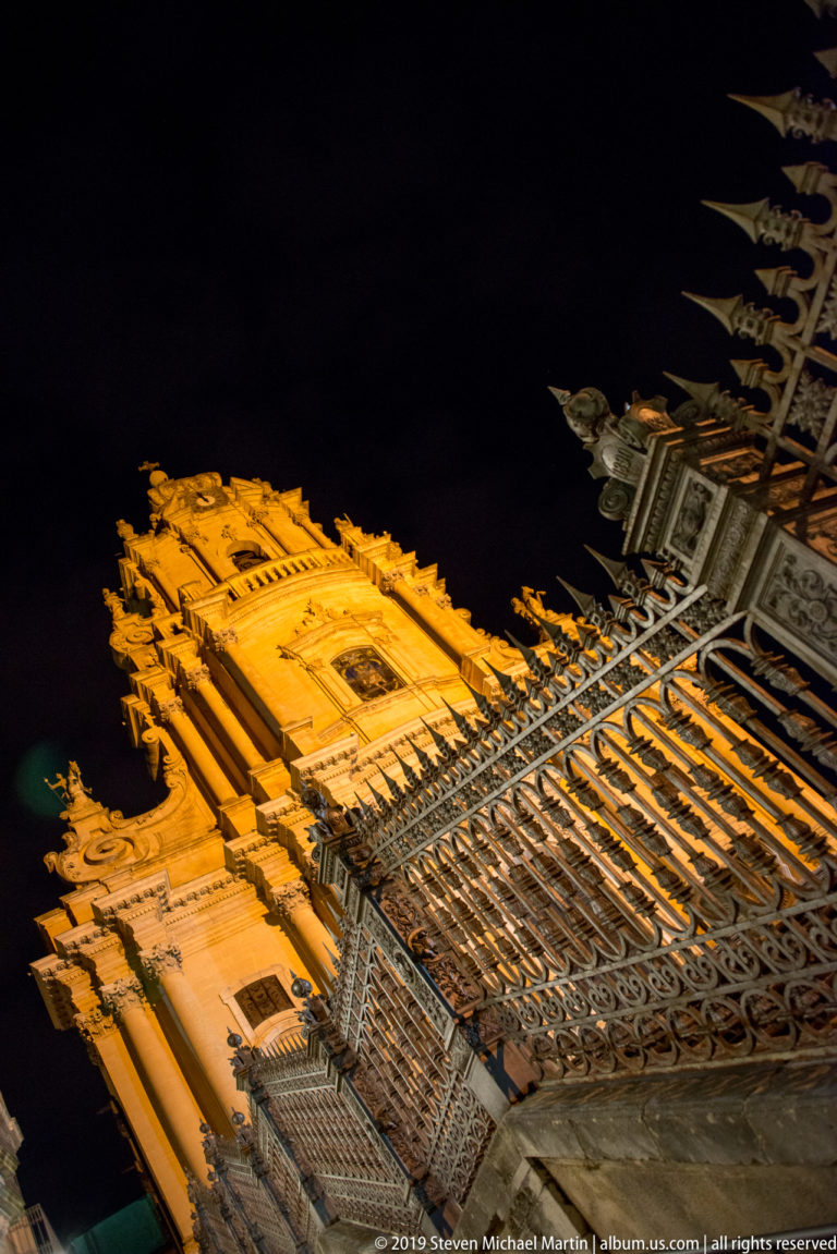 Steep low-angle nighttime view of an ornate Baroque church façade behind tall wrought-iron railings and pointed fence finials.