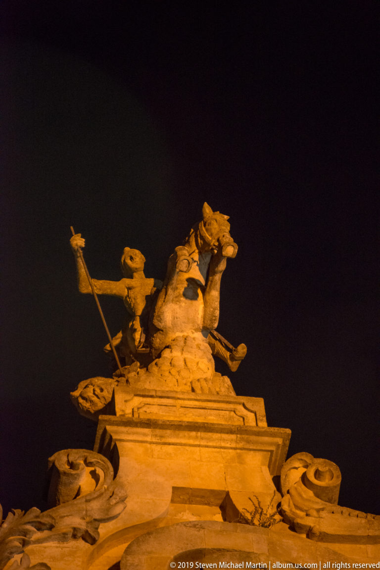 Upward nighttime view of a sculptural group with rearing horses and a chariot-like figure crowning an ornate Baroque façade.