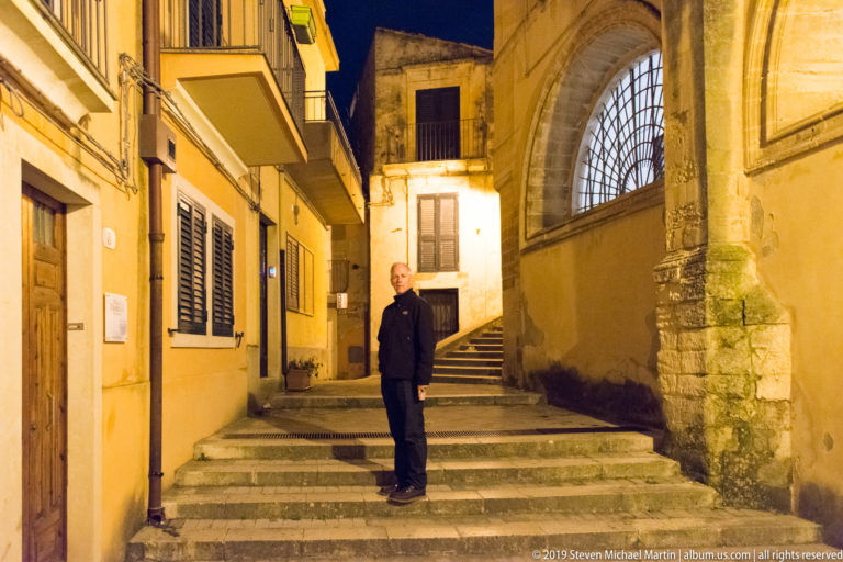 Man standing on broad stone steps between houses and a church wall with a tall arched, grilled window.