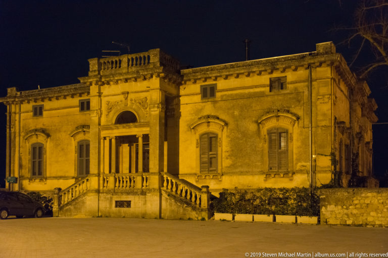 Frontal nighttime view of a symmetrical stone villa with twin balustraded stairs, shuttered windows, pilasters, and roofline balustrade.