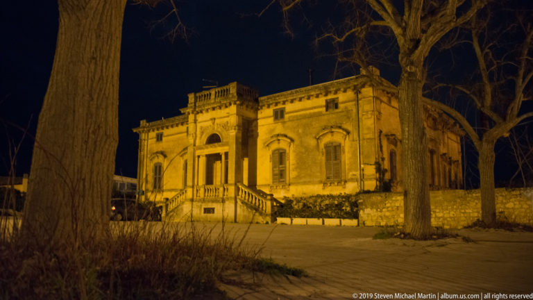 Illuminated historic villa with central balustraded staircase, symmetrical windows, and tall leafless trees in a paved forecourt.